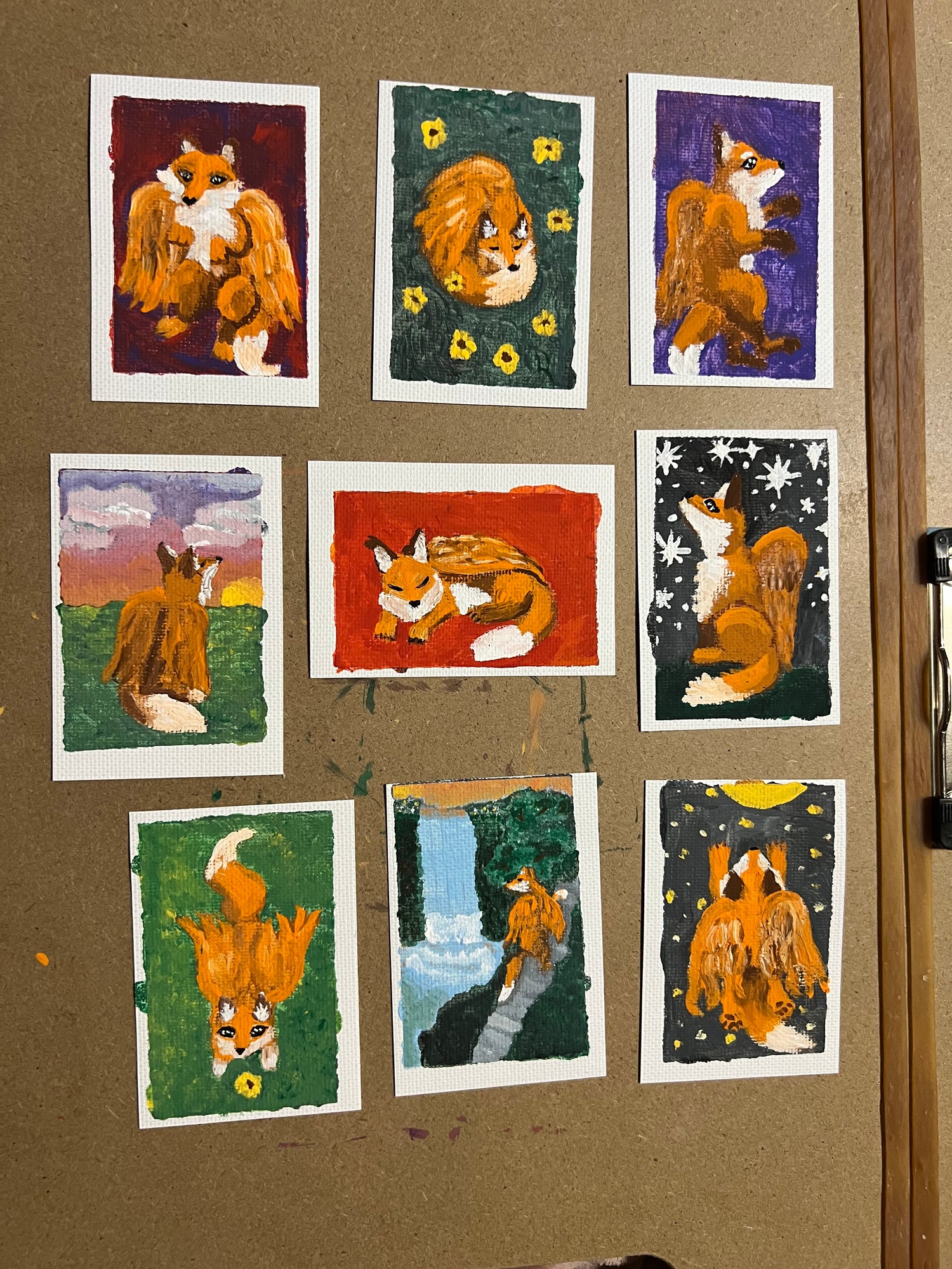 Nine small painted art cards featuring foxes in different scenes, arranged on a brown corkboard. The paintings use textured colors like orange, green, purple, red, and blue, with white borders around each card.