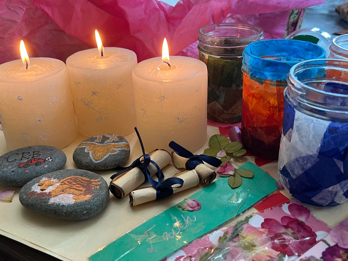 A styled tabletop display with four lit cream pillar candles, several painted stones, rolled parchment notes tied with blue ribbons, and multiple glass jars containing colorful dried flowers or petals, set against pink tissue paper.