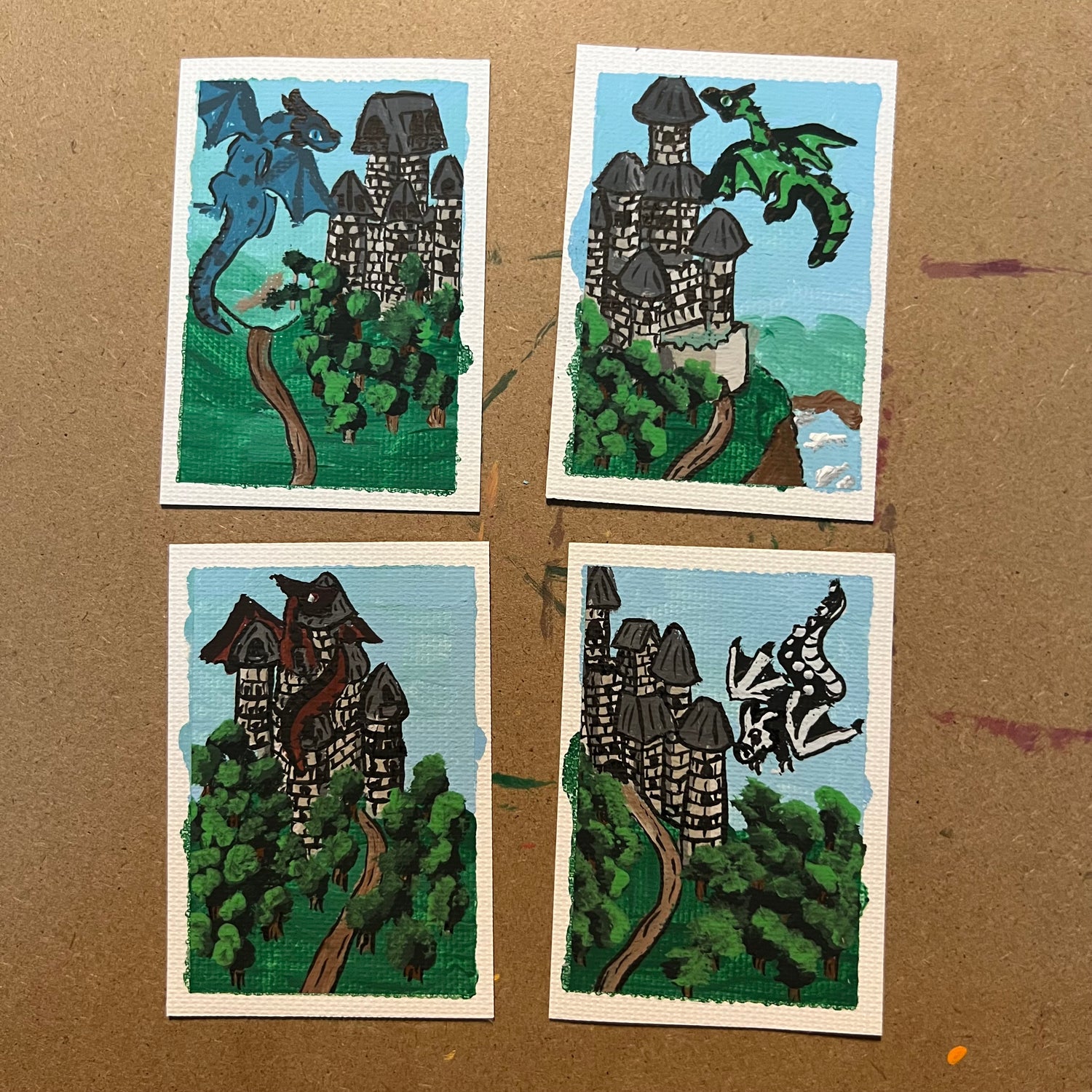 Four small illustrated cards or prints arranged in a 2x2 grid on a brown cardboard surface. Each card shows a fantasy castle scene with green trees, a winding path, blue sky, and a dragon in different poses and colors.