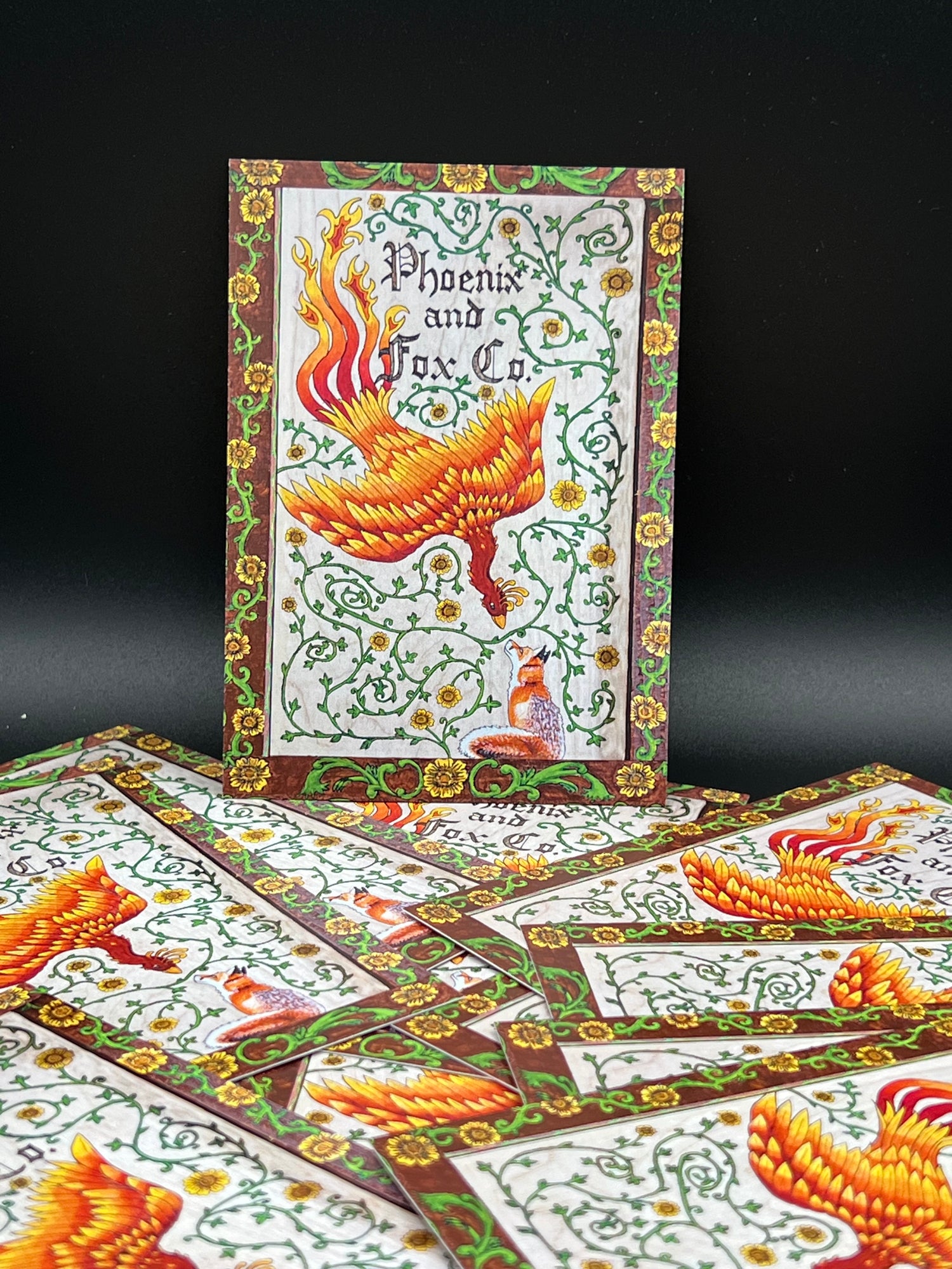 A deck of illustrated playing cards featuring a detailed phoenix design with green vines and gold floral borders on a white background, displayed with one card laid flat and others fanned out on a black surface.