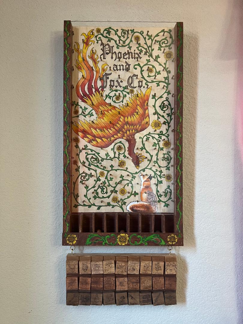 Image of a Plinko board that has a phoenix flying downward towards a fox looking up at it. Vinery throughout with yellow flowers. A brown border with vines and yellow flowers. A board hanging from the plink board with three rows of nine wooden tiles with small images on each tile.